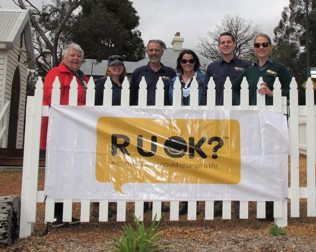 two men and 4 women in front of a white gate with banner